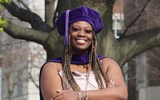 Woman in pink dress wearing a purple graduation cap standing in front of a campus building